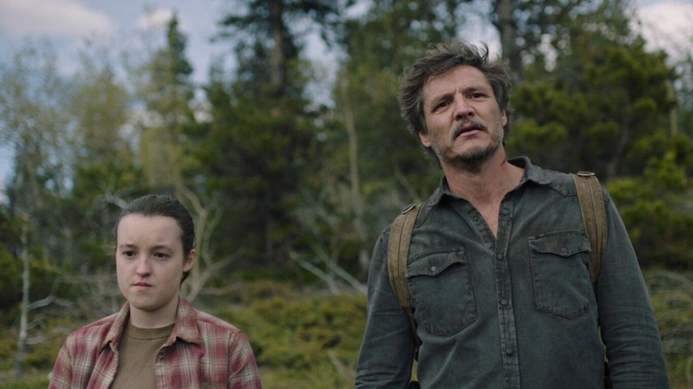 Ellie and Joel in the forest. Ellie, portrayed by Bella Ramsey, is a young white woman with dark hair and eyes. She wears a checked red and white shirt over a brown t-shirt. Joel played by Pedro Pascal is a tall white man with light brown and grey hair including a moustache and beard. He wears a blue shirt and a light brown backpack. Behind the two is the green trees and bushes of a forest. 