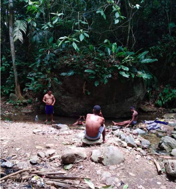 Four people with dark skin at rest in the jungle of the Darién Gap. In the foreground a man wearing white shorts sits on a rock with his back to the camera. To the left is a man wearing blue shorts standing with his left hand on his belly. In the center a man lies on his belly in a shallow pool of water. To the right a woman wearing a bikini top and shorts sits on a rock, facing side-on. Their spot is shaded by vivid green trees and bushes.