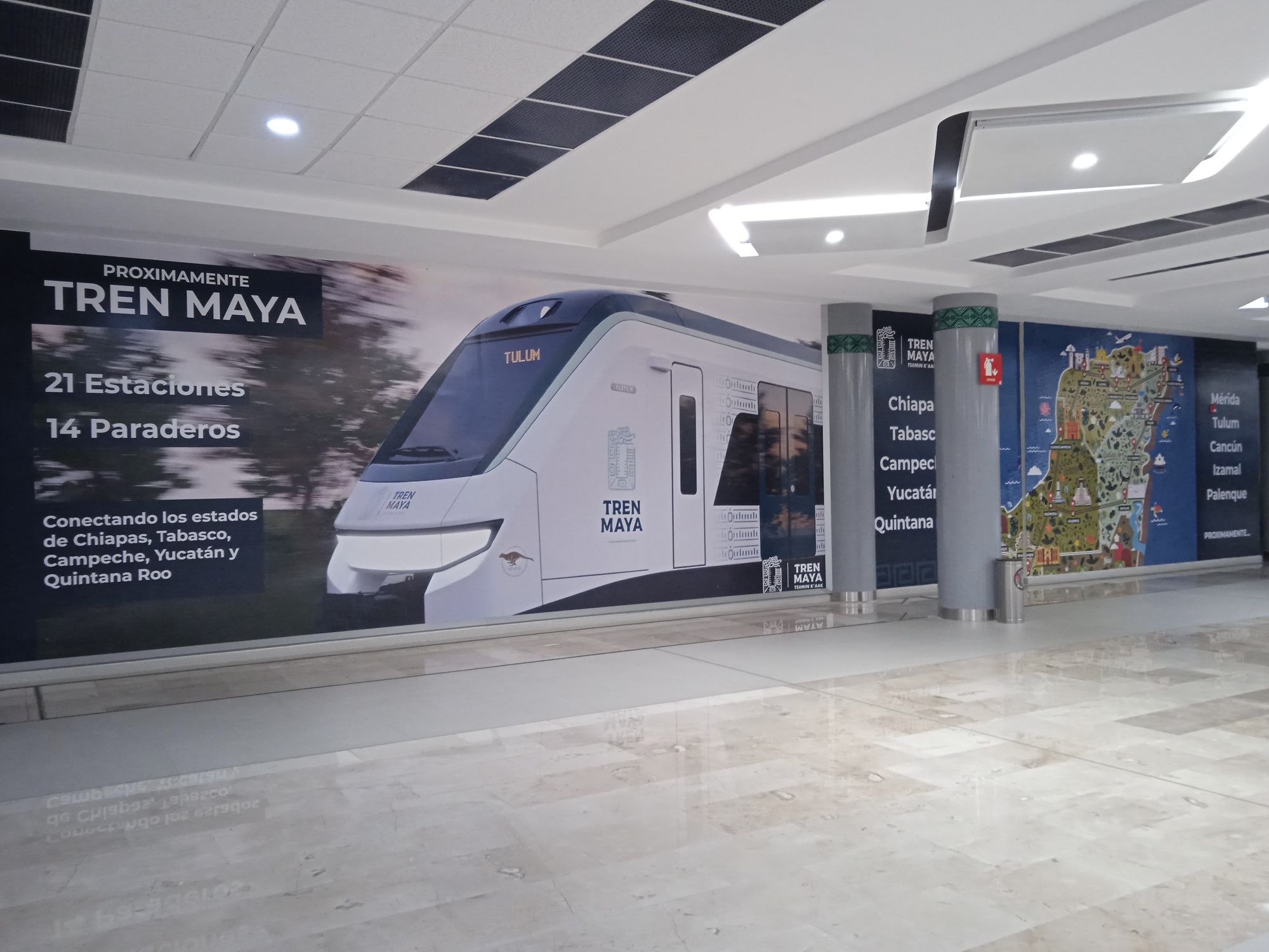 Baggage collection hall at Felipe Ángeles international airport, Estado de México. Pale beige flooring meets a wall covered in an advertisement for the Tren Maya. The ad shows a picture ofo an electric train - a blue and white front carriage with the destination 'TULUM' on the front up the top in digital letters. On the left, large white lettering says: Proximamente Tren Maya (Maya Tren Coming Soon), 21 Estaciones 14 Paraderos (21 stations 14 stops), Conectando los estados de Chiapas, Tabasco, Campeche, Yucatán y Quintana Roo (Connecting the states of Chiapas, Tabasco, Campeche, Yucatán, Quintana Roo). On the right the viewer can glimpse a map of the train's route in a series of green, blue and yellow tones. Photo: Ann Deslandes, August 2023