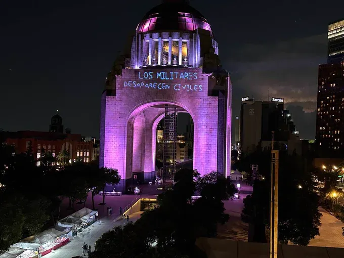Mexico's Monument to the Revolution, consisting of a four-legged edifice made of brick with a dome on the top, is lit up in purple. Underneath the dome is the words 'Los militares desaparecen civiles'/'The military disappears civilians', in white projected lettering. The purple edifice is lit up against a dark sky and surrounded by city buildings that are also dark.